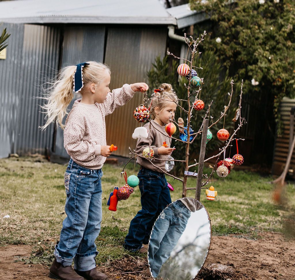 Two children decorating a small tree with colorful ornaments outdoors.